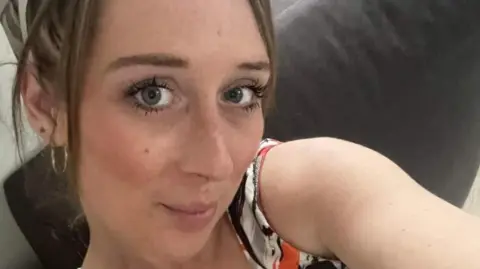 Family Handout/PA Alana Odysseos is taking a selfie wearing gold hoop earrings and an orange, white, grey and black patterned sleeveless top. She is sitting on a grey sofa.