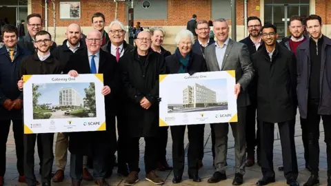 A large group of people stand outside Doncaster Railway Station holding up boards showing architectural plans for a new office building.