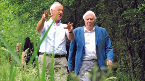 Tom Marshall Sir David Attenborough and Alastair Fitter walking together along a narrow, grassy path in a lush, green woodland area.