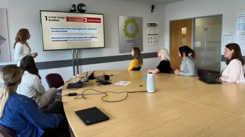 A group of women in a boardroom sat around a table looking at a presentation. On the screen the words "Avoid Victim Blaming' can be seen.