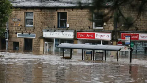 A bus stop and a row of shops under water in Hebden Bridge