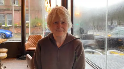 A woman with blonde hair and a green top sitting on front of a large window. She has a slight smile on her face and rain coats the windows next to her.