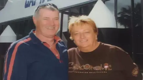 A man in a polo shirt and a woman in a t-shirt. Both are looking at the camera and smiling. They have an arm around each other.
