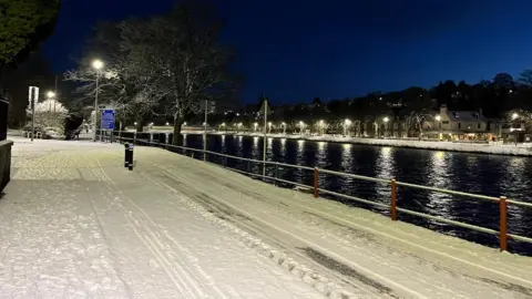 Snowfall on a road in Inverness. It is early morning and dark and the street lights are on.