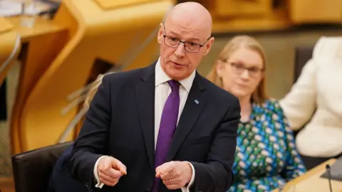 PA Media John Swinney, who is bald with glasses, in the Scottish Parliament chamber. He is wearing a dark suit, white shirt and purple tie. 