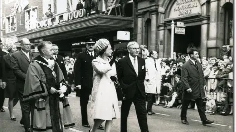 HANCOCK & WOOD Black and white shot of the Queen walking past Hancock and Wood in 1968. She is waving and walking in the middle of the road with a long crowd behind her.
