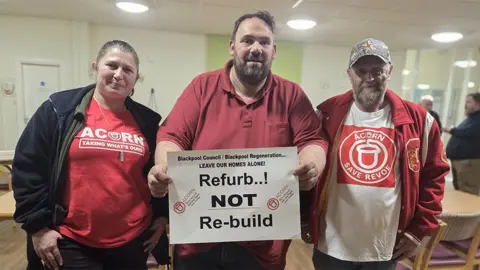 Residents Anna Penfold, John Dacks and Michael Edge hold a poster in the community centre that says "refurb not rebuild"