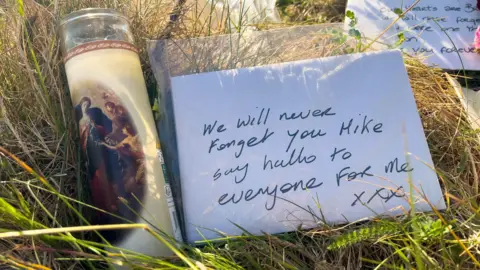 PA Media A piece of white paper with black font reading 'we will never forget you Mike say hello to everyone for me xxx' is enclosed inside a transparent cover. Beside the piece of paper is a cream candle with a religious picture on it. Both are sitting on grass. Another piece of paper can be seen in the background.