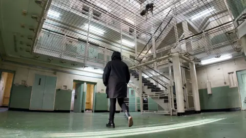 BBC Ben Moore A former prison worker walking the landings of a former prison in Kent. He is wearing and dark hooded coat and is viewed from behind. The prison has a green floor, is stark and empty, with metal staircases and thick doors.