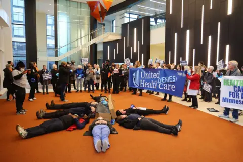 Getty Images Protesters from the Popular Action Movement lie in a circle in front of the headquarters of health insurance giant UnitedHealth Group, holding signs reading "UnitedHealth denies sponsorship"