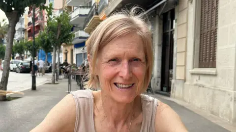 Di Gomm Di Gomm has light blond hair and is wearing a vest and a necklace. She is on a table outside in the sun. She is smiling. 