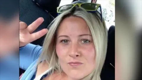 Joanne Penney looks straight at the camera, she has blond hair and is holding up her right hand with a v sign. She is not smiling.