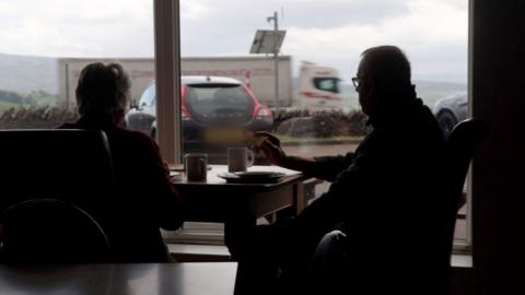Two people sit and look out of a window at a passing lorry