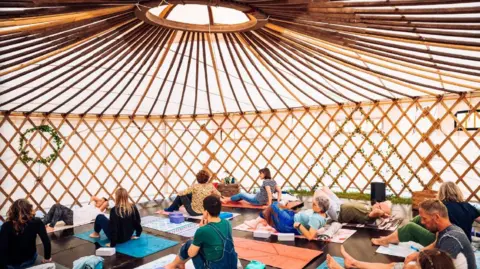 Fire In Your Soul People are sitting on yoga mats in a yurt. They are doing yoga exercises that a teacher in the front is demonstrating.