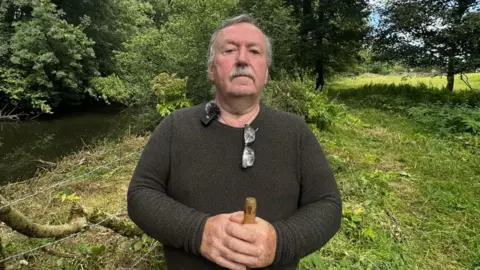 A man with short grey hair, a moustache, glasses around his neck and a brown jumper. He holds a brown wooden stick in his hand 