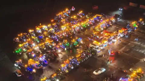 David and Obby Stevenson Aerial view of tractors, vans and farming equipment lined up in rows at night time in a car park - around 60 or so - all brightly lit with Christmas lights as people walk around each vehicle