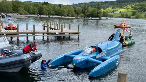 Bluebird is towed on to the shore by a support crew in a small boat. A woman is in the water in front of the craft while another woman leans over a support boat. Coniston's fells can be seen behind.