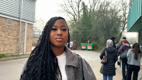 A young woman is standing next to a queue of people outside a university building, She is wearing a white top and a grey jacket.