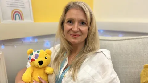 A woman with long blonde hair and blue eyes smiles directly at the camera. She is wearing a white shirt and has a blue lanyard on. She is holding a small Pudsey Bear toy and sat on a cream coloured sofa. In the background - fairy lights and posters can be seen - one has a rainbow on it.