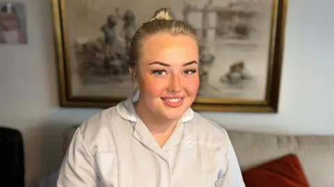 Supplied A woman in her nurse's uniform smiling at the camera. Behind her is a sofa with a red cushion, and a picture on the wall.