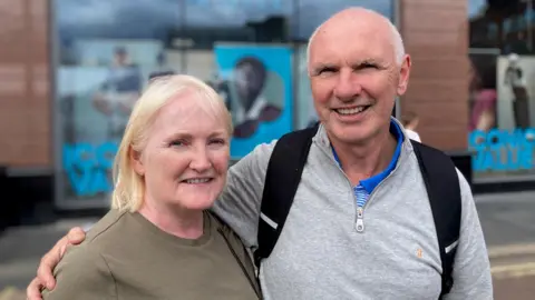 A woman and a man smile at the camera. The woman has blonde hair and is wearing a pale-green top. The man is wearing a grey top. 