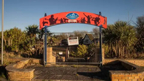 Alfie Lugsden/Pleasure Island Archive Exterior view of the former entrance to the park. There is a red sign above the entrance, which has been cordoned off with a metal fence panel. There are a number of cordyline-type plants close to the entrance and some buildings in the distance. 
