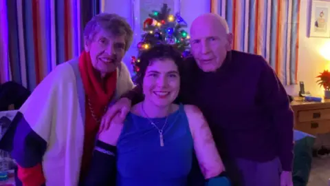 Lily McGarry Lily and her grandparents are posing in front of a small, decorated Christmas tree with colorful lights. The room has striped curtains, a framed picture on the wall, and warm ambient lighting.