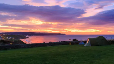 Two tents are pitched on a patch of grass overlooking Whitby. The sky is pink with a sunset.