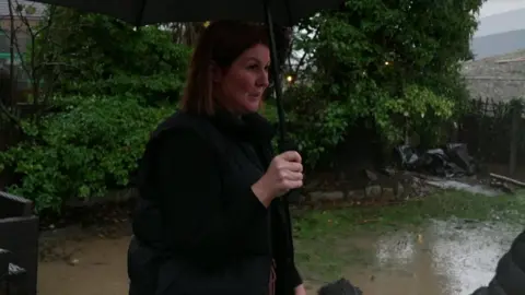 Paula stands in her back garden holding a black umbrella. A large pool of brown water covers the grass around her.