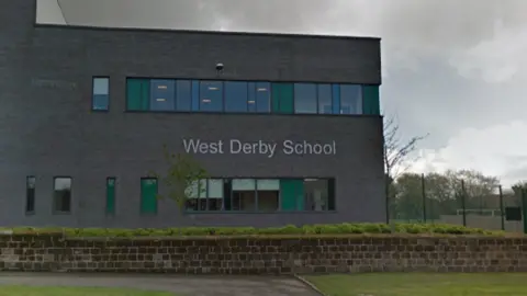 A grey block building with lettering spelling West Derby School on the front stands behind a low brick wall surrounded by wedges. In front of the wall are two strips of grass and a concrete path. There are grey clouds in the sky.
