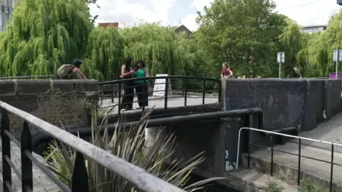 People stand and walk on a footbridge over a canal in a leafy urban area, with weeping willows and buildings are seen in the background