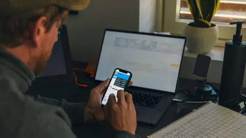 Getty Images File image of a man in a grey top looking at data on his mobile phone while a notebook with a hand-drawn table sits on a table to his right, and a blurred-out screen on a laptop can be seen in the background