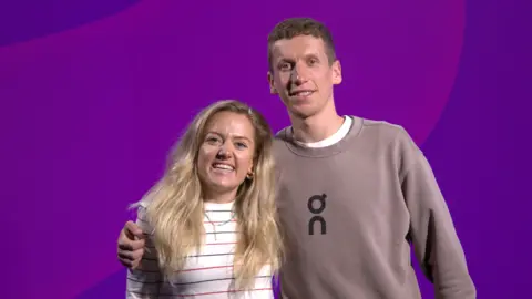 Anya Culling has long blonde hair and is wearing a white sweater with red and blue stripes as she smiles for the camera with Logan Smith to her right. He is taller and standing with his arm around Anya. He is clean shaven, has short brown hair and is wearing a brown jumper over a white t-shirt. They are standing in front of a screen with which is showing a two-tone purple pattern.