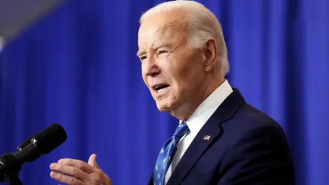 Reuters Joe Biden stands side-eye on to the camera, and gesticulates in front of a microphone against a blue curtained backdrop. He wears a navy blazer and white and blue tie. 