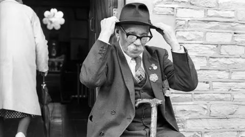 A black and white photograph of an old man with a white moustache rearranging his Trilby hat 