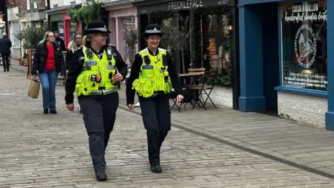 Two uniformed police officers on patrol in Lincoln city centre. A number of businesses can be seen in the background, along with people out shopping.