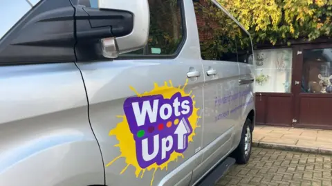 Vikki Irwin/BBC A silver van is parked on a paved area next to a building with wooden panels and glass windows. The van has a colourful logo on the side that reads WOTS UP in bold white letters on a purple background with a yellow splash design behind it. 