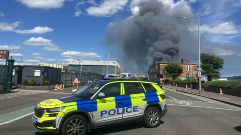 Yellow, blue and white police vehicle at the scene of a fire in Belfast at East twin Road. There is black smoke and a brown brick building in the distance.