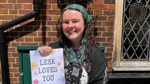 A lady with a green bandana on her head. She has long black hair with green streaks going through it. She is wearing a black top and holding up a paper sign that says "Leek loves you".