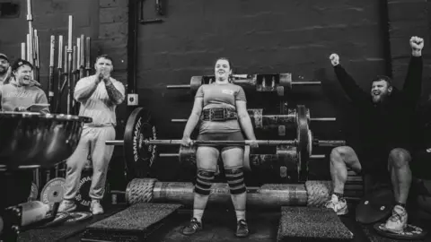 Gordon Siegal A black‑and‑white gym scene shows Lauri in the centre performing a heavy deadlift on a platform surrounded by weights. Several individuals, including the Stoltman brothers, stand nearby clapping and raising their arms, positioned along the sides of the lifting area. Barbells, plates, and gym equipment line the background against a dark wall.