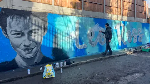 A man touches a wall on which a gigantic blue mural has been painted. On the left is Leo Ross's face, neck, and shoulders. On the right is his name, painted on the wall in white writing.
