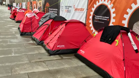 A line of red tents outside Manchester town hall, pitched on wet stone paving, under some fencing covered in advertising.