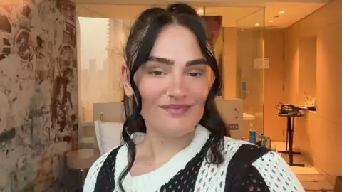  Tiegan McFadden, who has, dark hair looking at the camera, she is wearing a black and white knitted cardigan and is standing in a hotel room. An ironing board and iron can be seen in the background.