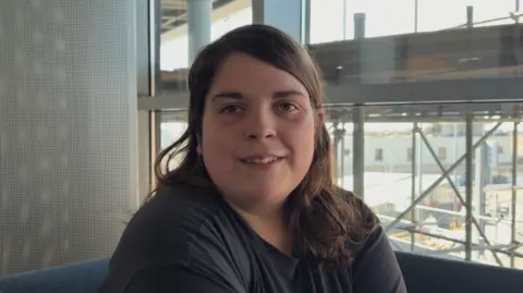 Chloe Browning is a young woman with long brown hair. She is sat down and wearing a dark T-shirt while smiling at the camera.