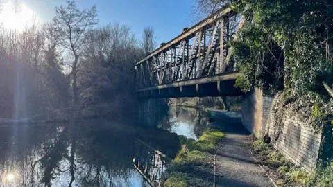A steel truss bridge over a canal in Wolverhampton on a bright day, with a blue sky and a large flare of light in the top-left corner of the photo. Trees line the left-hand side of the canal and are reflected on the water. There is a footpath on the right side.
