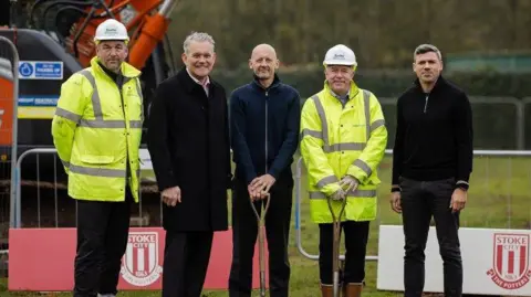 five men - two in high visibility jackets, the rest in dark-coloured clothing - stand with construction machinery and Stoke City branded boards behind them. Two of the men are holding shovels.