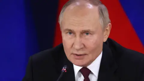 Getty Images Vladimir Putin, wearing a black suit and red tie, speaks into a small black microphone.