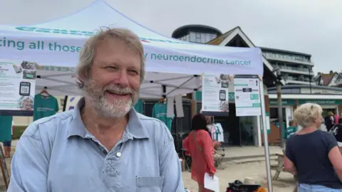 A picture of Dave smiling in front of a large white tent. He is wearing a blue polo shirt.