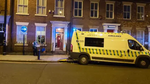 An ambulance is parked outside a terraced house in Sunderland. A man stands outside wearing a mask.