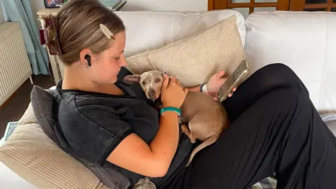 Clare Phillips Tilly, is sat on a couch with a small dog on her lap. She is looking at a phone which she is holding. 